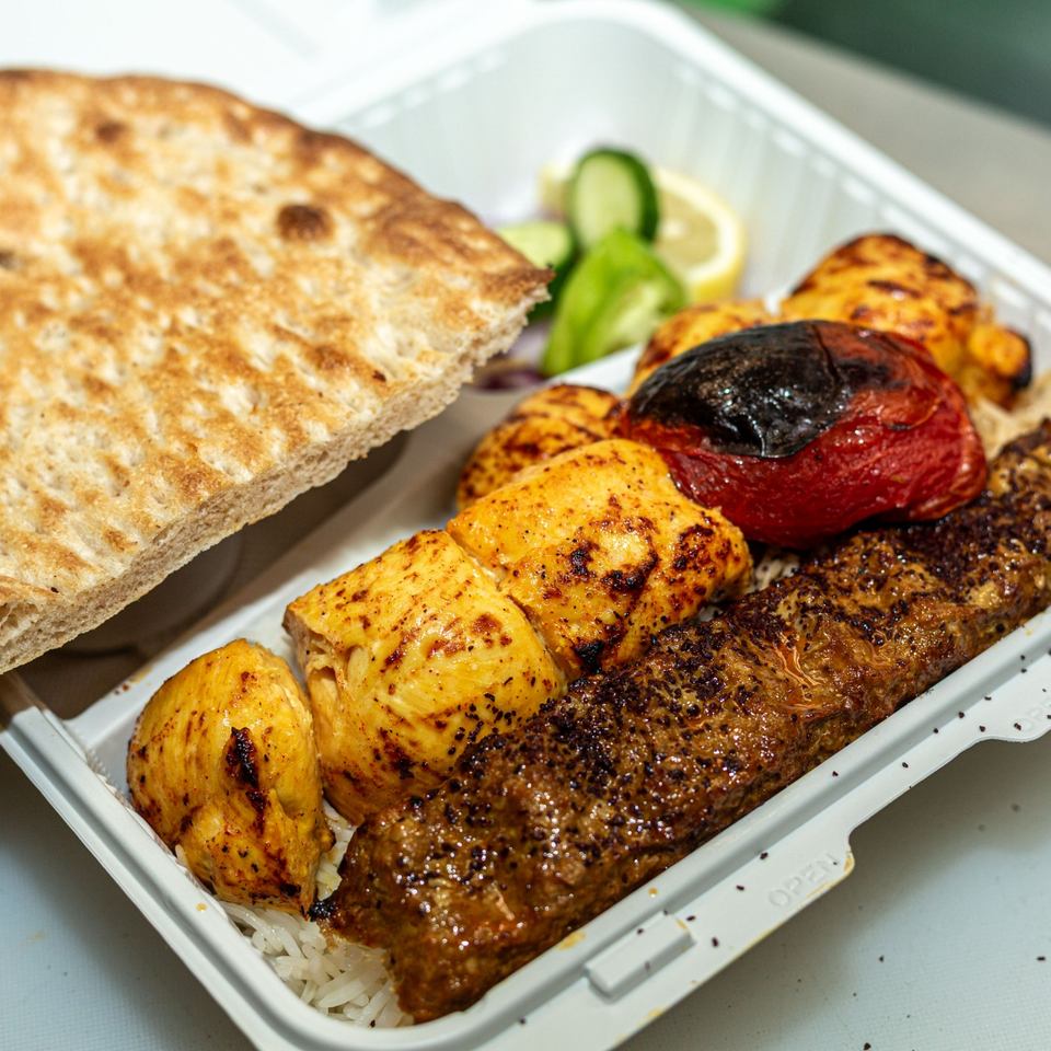 Grilled chicken, beef kebab, roasted vegetables, rice, flatbread, and salad arranged in a takeaway tray.